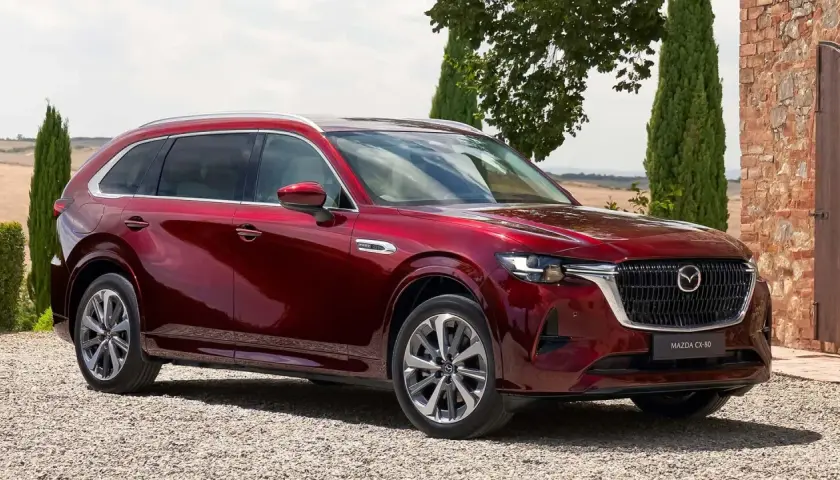 Lśniąca, czerwona Mazda CX-80 zaparkowana na żwirowej drodze, otoczona zielenią i ceglanym murem.