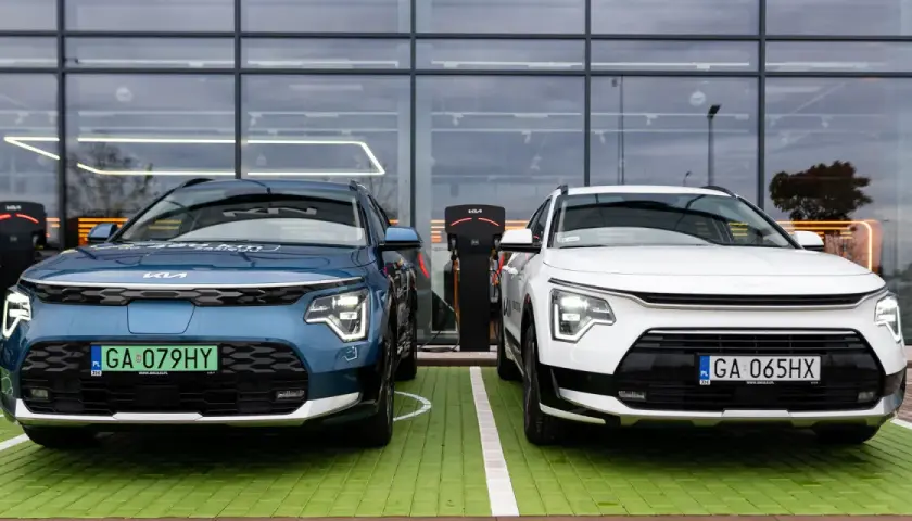 Dwa nowe samochody Kia Niro, jeden niebieski, drugi biały, stoją obok siebie na zielonym parkingu przed nowoczesnym budynkiem z dużymi oknami.