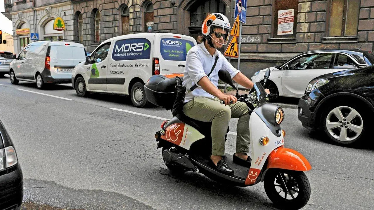 Mężczyzna w kasku jedzie skuterem elektrycznym, posiadając prawo jazdy AM. W tle białe vany dostawcze.