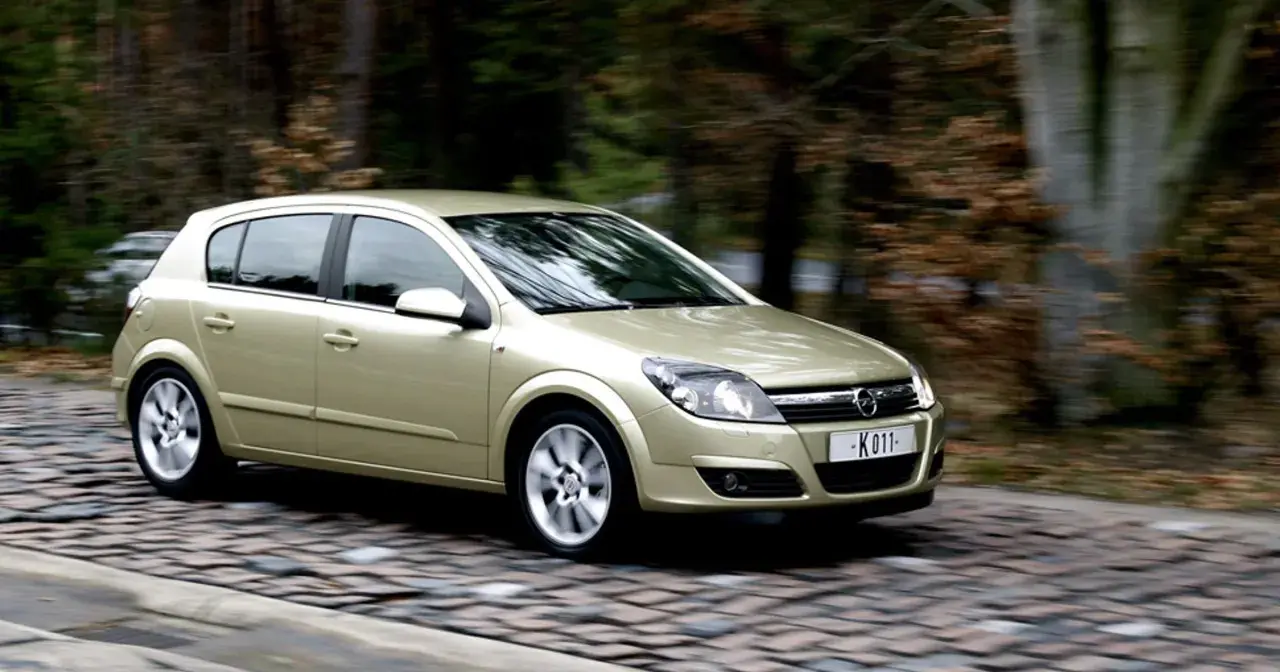 Złoty Opel Astra H 1.6 benzyna w ruchu na brukowanej drodze, otoczony jesiennym lasem.