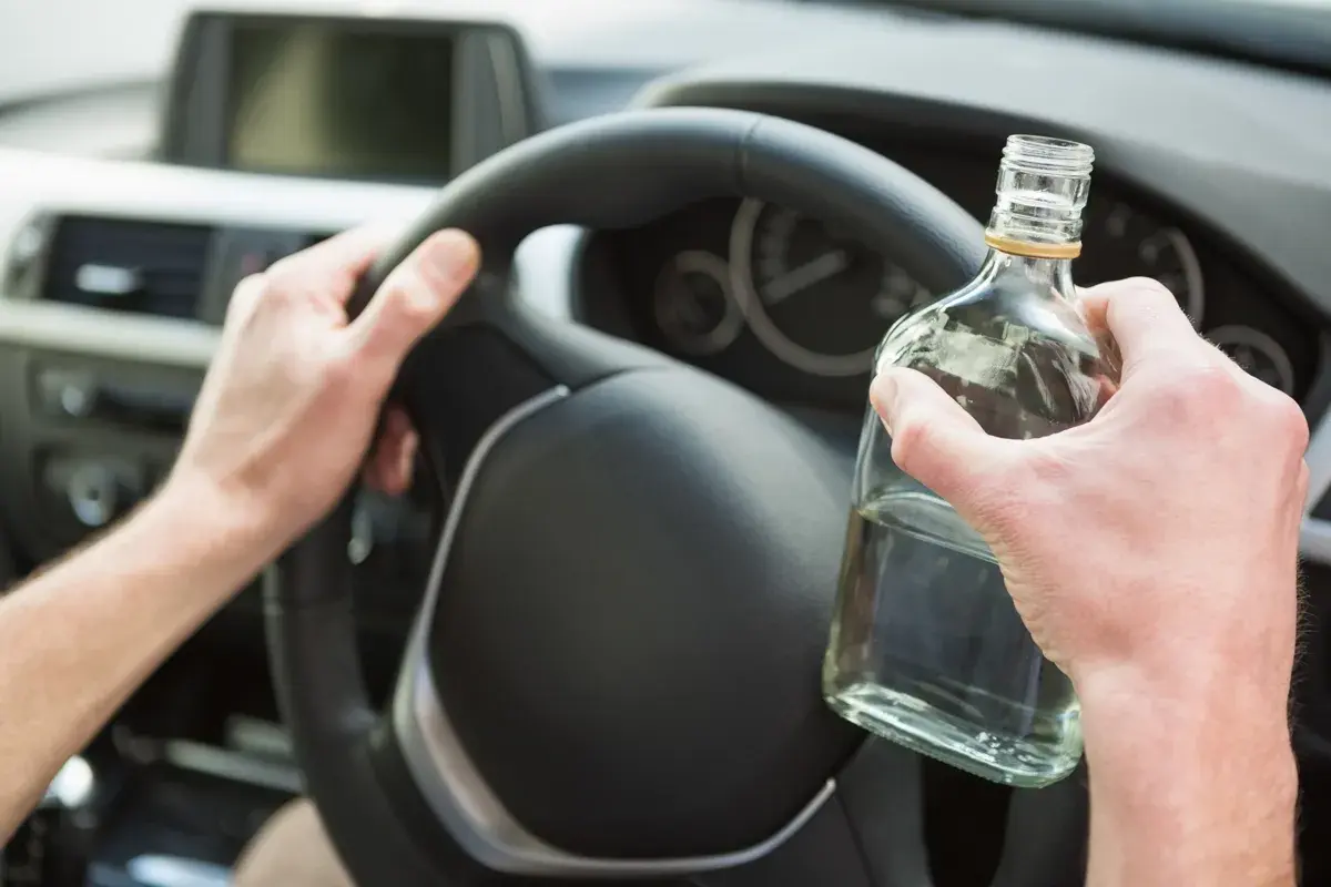 Ręce kierowcy na kierownicy, jedna trzyma butelkę alkoholu. Nie warto sprawdzać, ile promili.
