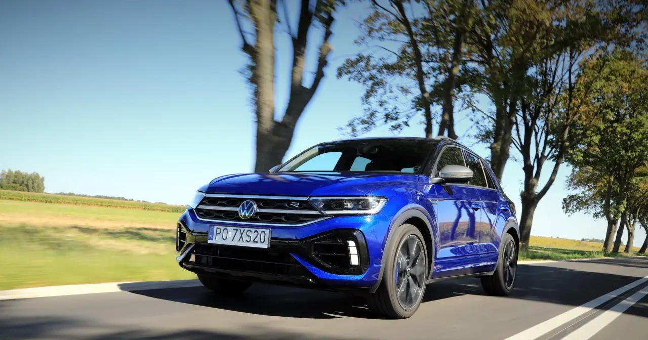 Niebieski Volkswagen T-Roc R pędzi po wiejskiej drodze, otoczony drzewami i polami.