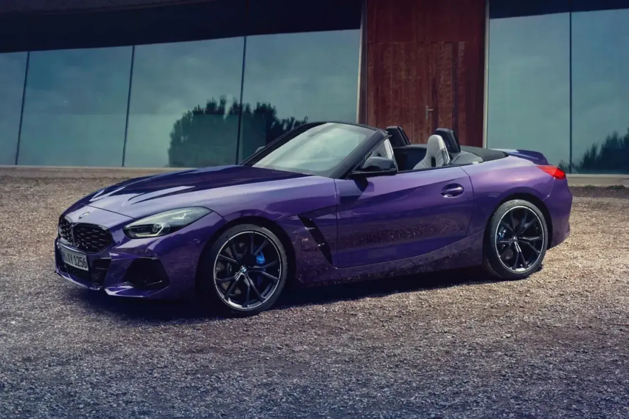 Fioletowy roadster BMW Z4 z otwartym dachem, zaparkowany na żwirowej nawierzchni przed nowoczesnym budynkiem.