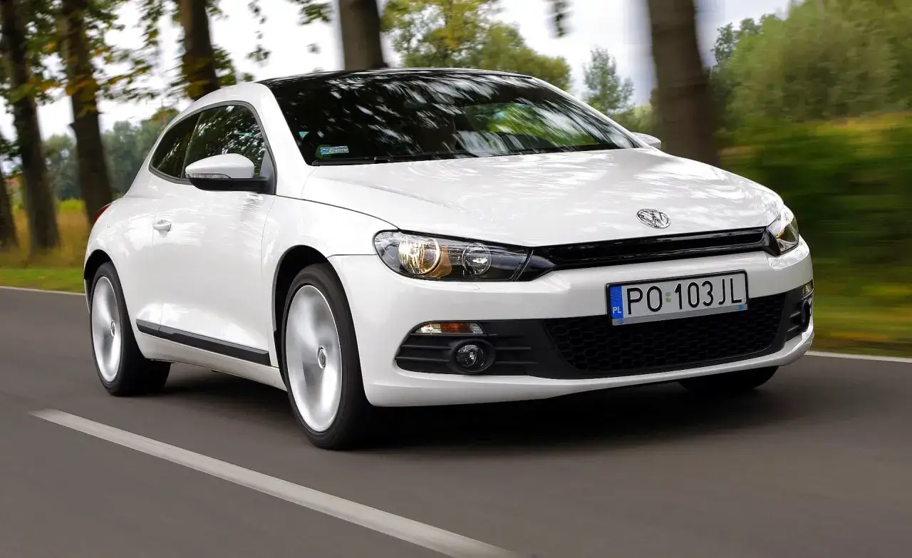 Biały Volkswagen Scirocco pędzi po drodze wśród drzew.