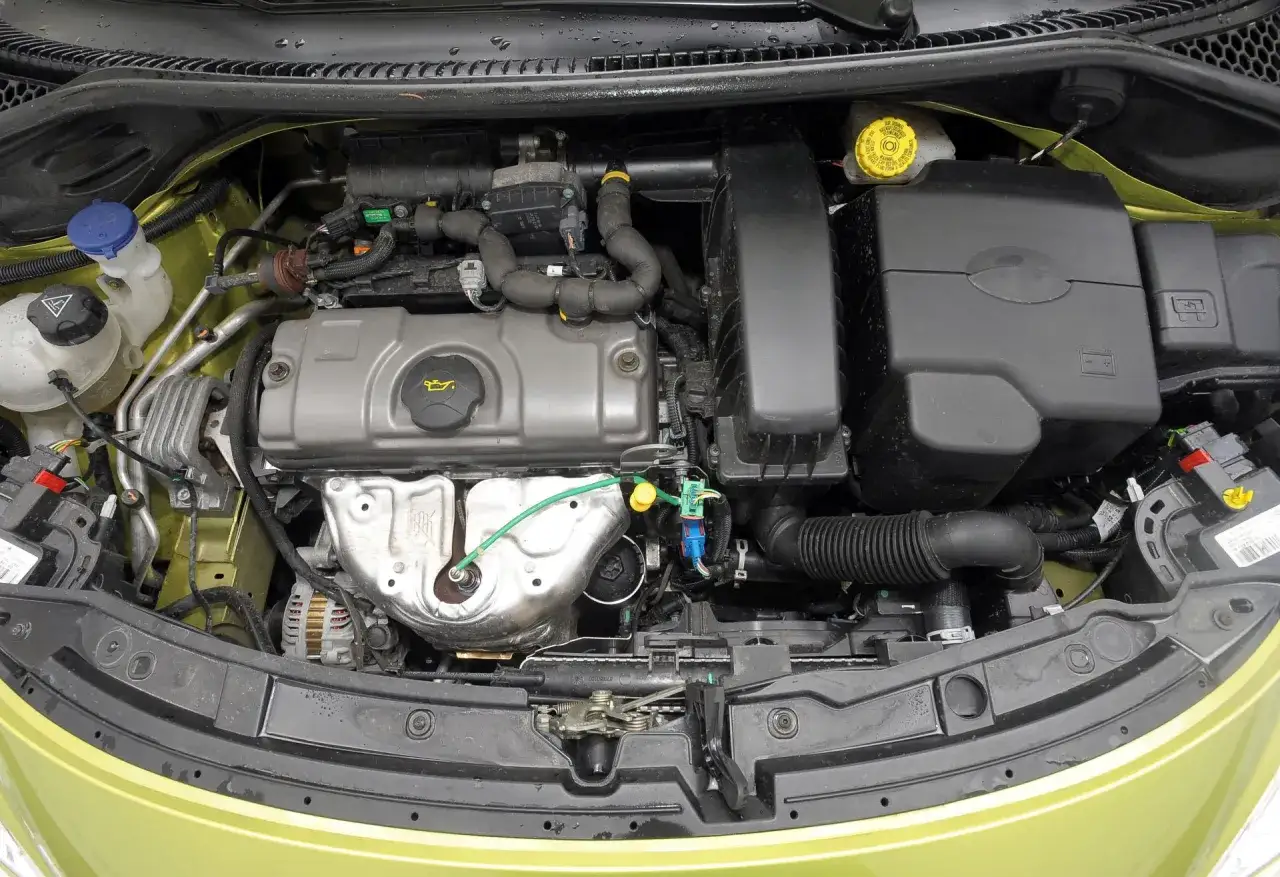 Silnik peugeot 207 1.4 benzyna. Widok komory silnika z widocznymi elementami, takimi jak filtr powietrza, zbiorniczek płynu chłodniczego i alternator.