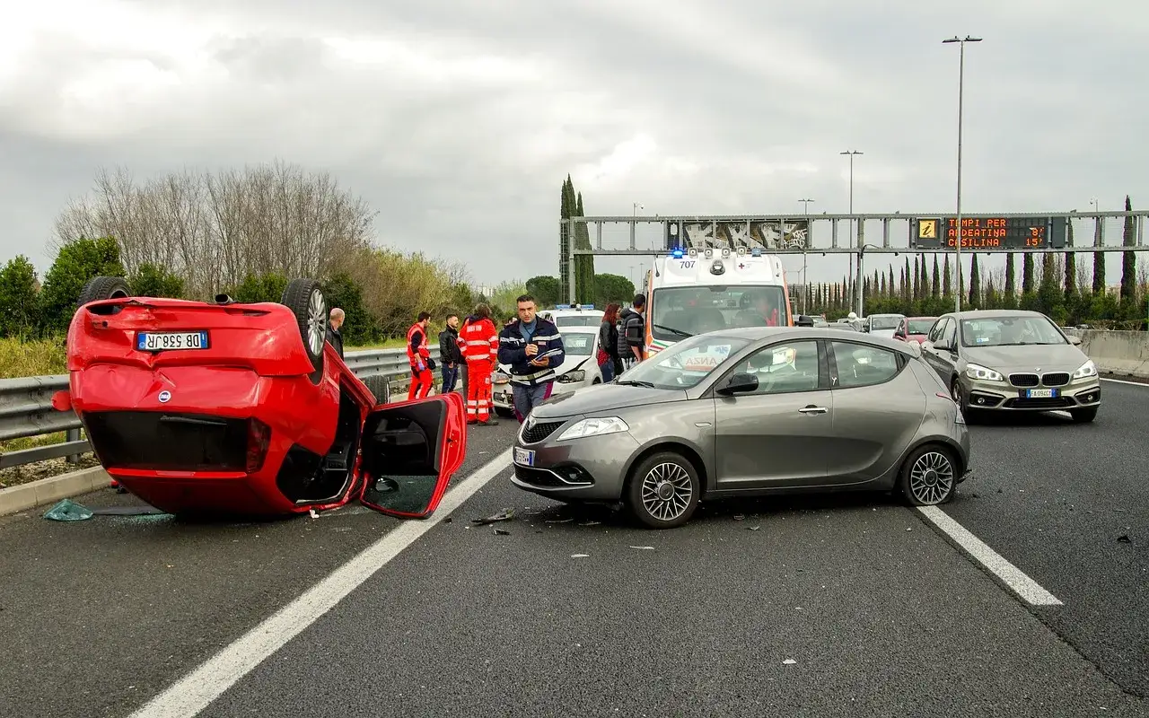 Na autostradzie doszło do poważnej kolizji drogowej. Czerwony samochód leży na dachu, obok stoi szary pojazd, a w tle widać karetkę i ratowników.