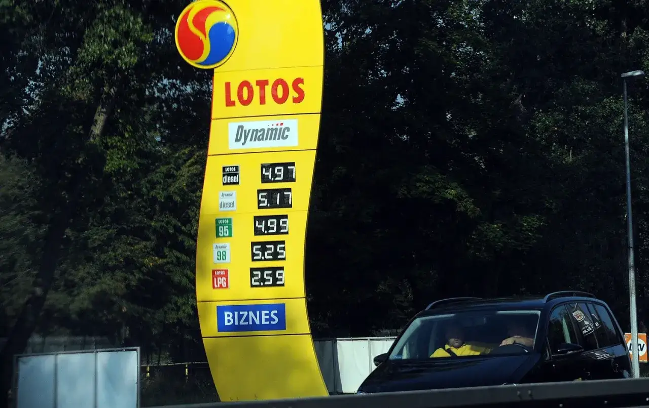 Ceny paliw na stacji Lotos: diesel 4.97, 95 4.99, LPG 2.59. Kierowca w żółtej kurtce obserwuje hurtowe ceny paliw Lotos.