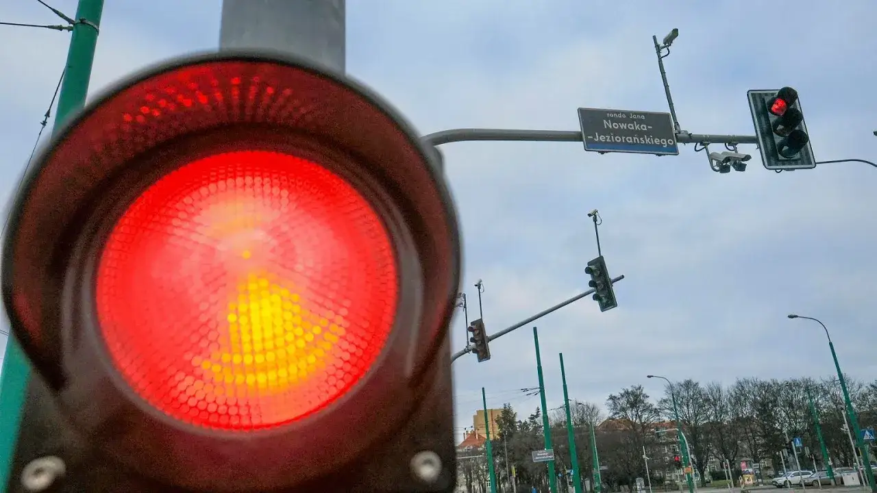Czerwone światło na sygnalizatorze drogowym, ostrzegające przed mandatem za czerwone światło. W tle znak z nazwą ronda.